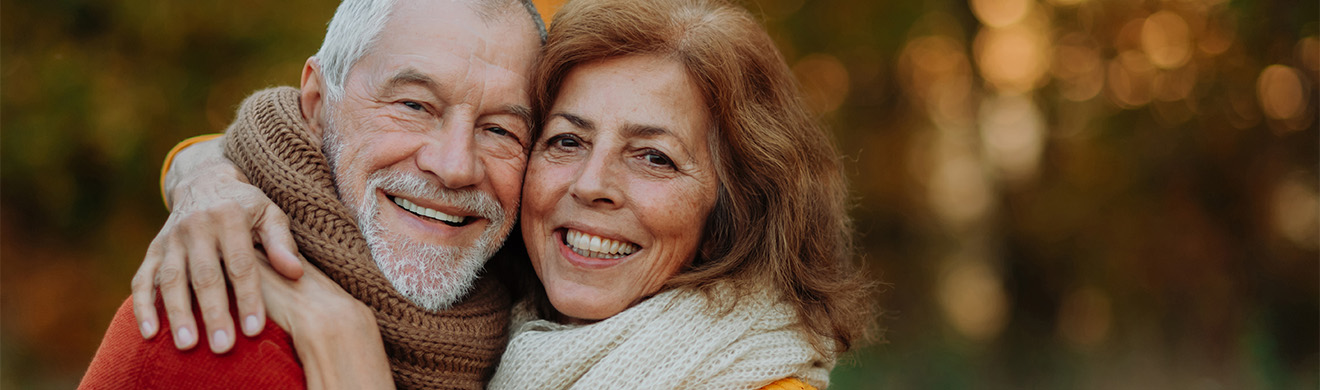 Seniorenpaar lächelt glücklich im Herbstwald, zeigt gesunde Zähne und natürliche Ausstrahlung mit ästhetischem Zahnersatz in Lüdenscheid.