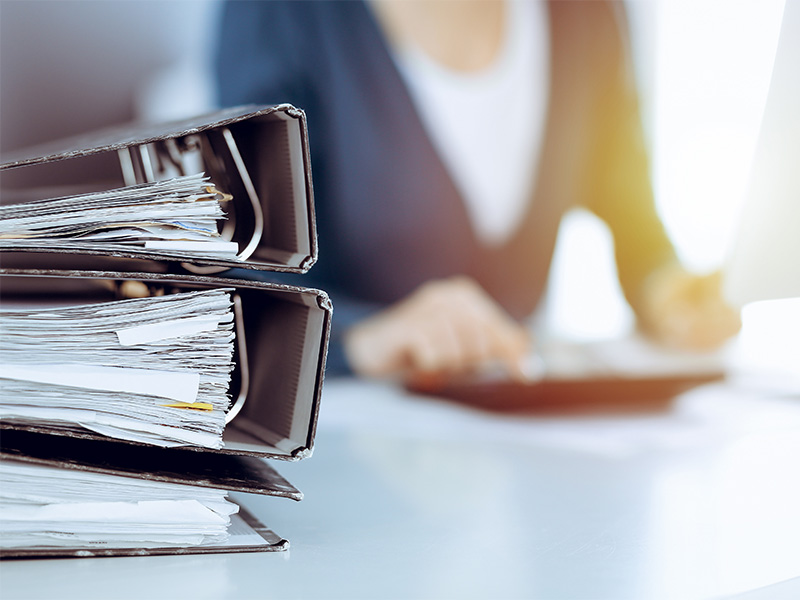 Ordner mit Geschäftsunterlagen auf Schreibtisch, im Hintergrund arbeitet eine Mitarbeiterin eines Wirtschaftsprüfers an Daten.